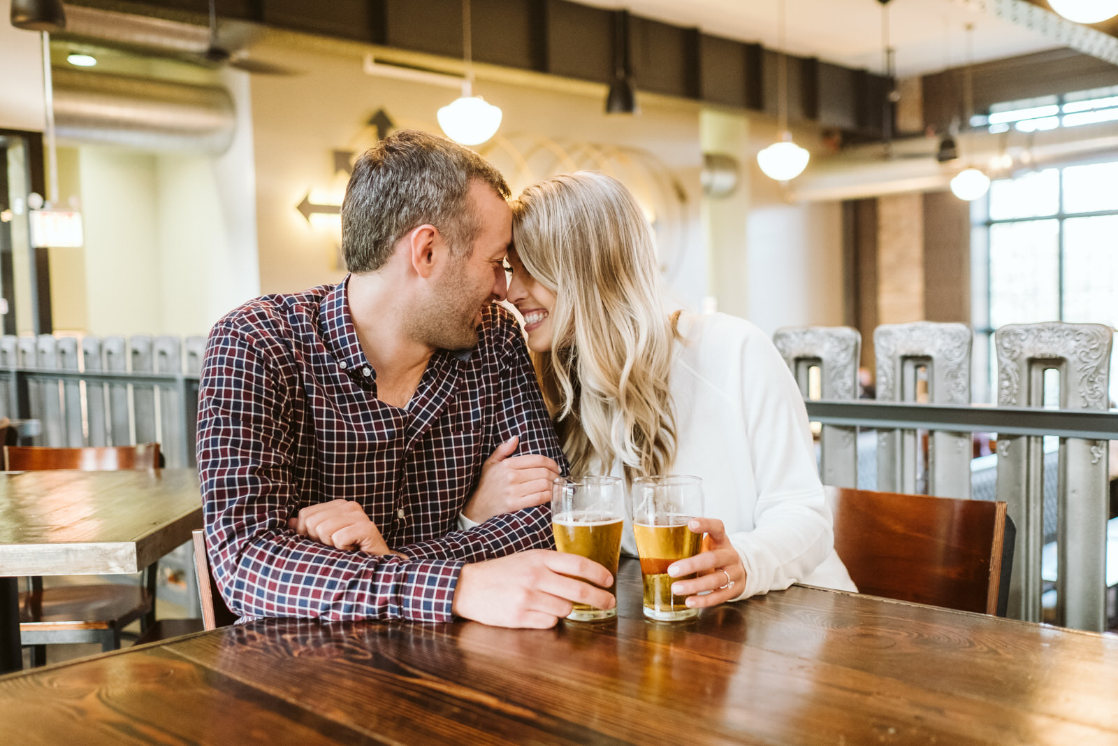 Couple cuddles at a hightop table while drinking beer for Chicago engagement session locations. Photography by Michael & Kristin Photo and Video.